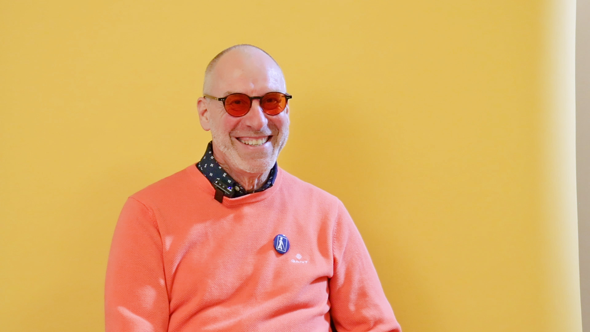 Man i medelåldern med solglasögon och orange tröja med synskademärke på skrattar.