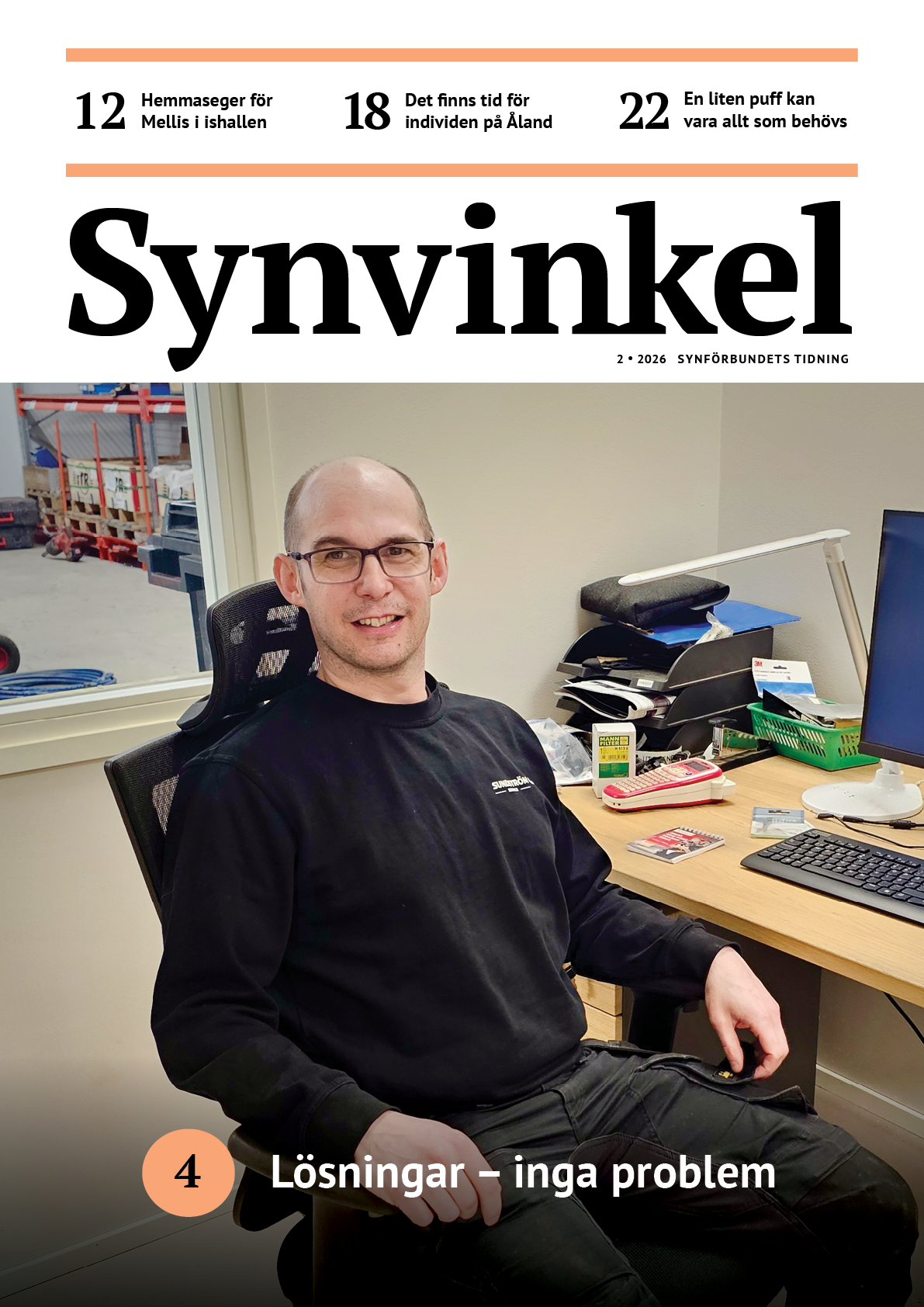 En leende, medelålders man med glasögon sitter i en kontorsstol vid ett skrivbord. Han är klädd i svarta arbetsbyxor och en svart långärmad tröja med texten Sundström på bröstet. Genom ett fönster bakom honom syns ett lager med röda hyllor.