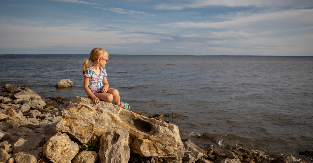 En liten flicka med ponnysvans och solglasögon sitter på en sten och blickar ut över ett öppet hav.
