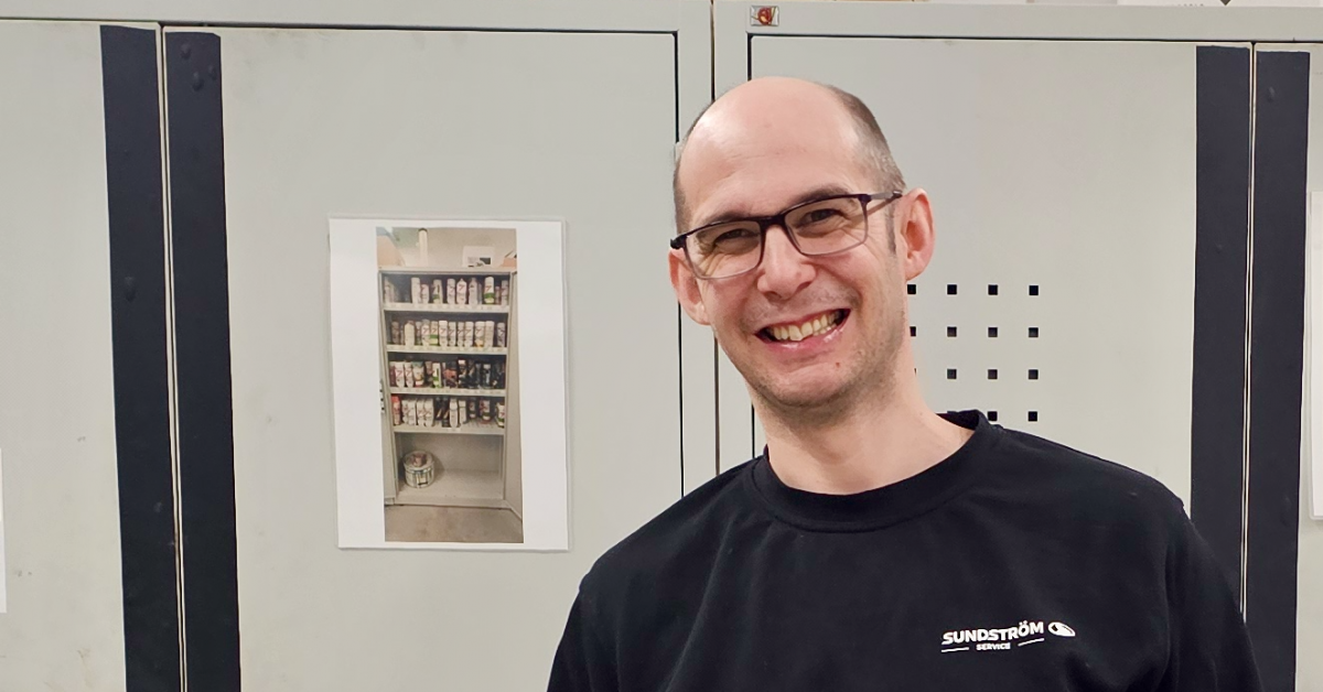 En leende man med glasögon står i arbetskläder framför gråa metallskåp i en lagerlokal. Flera av skåpen har svarta lodräta tejpremsor fästa på kanterna för att markera dem tydligare.