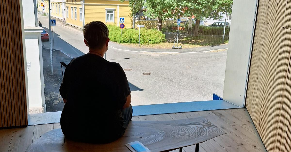 En person med kort hår sitter på en bänk framför ett stort fönster med utsikt över en gata och flera trähus i gul färg. Rummet är inrett i trä med både väggar och golv i samma träton, vilket skapar en varm känsla i interiören.