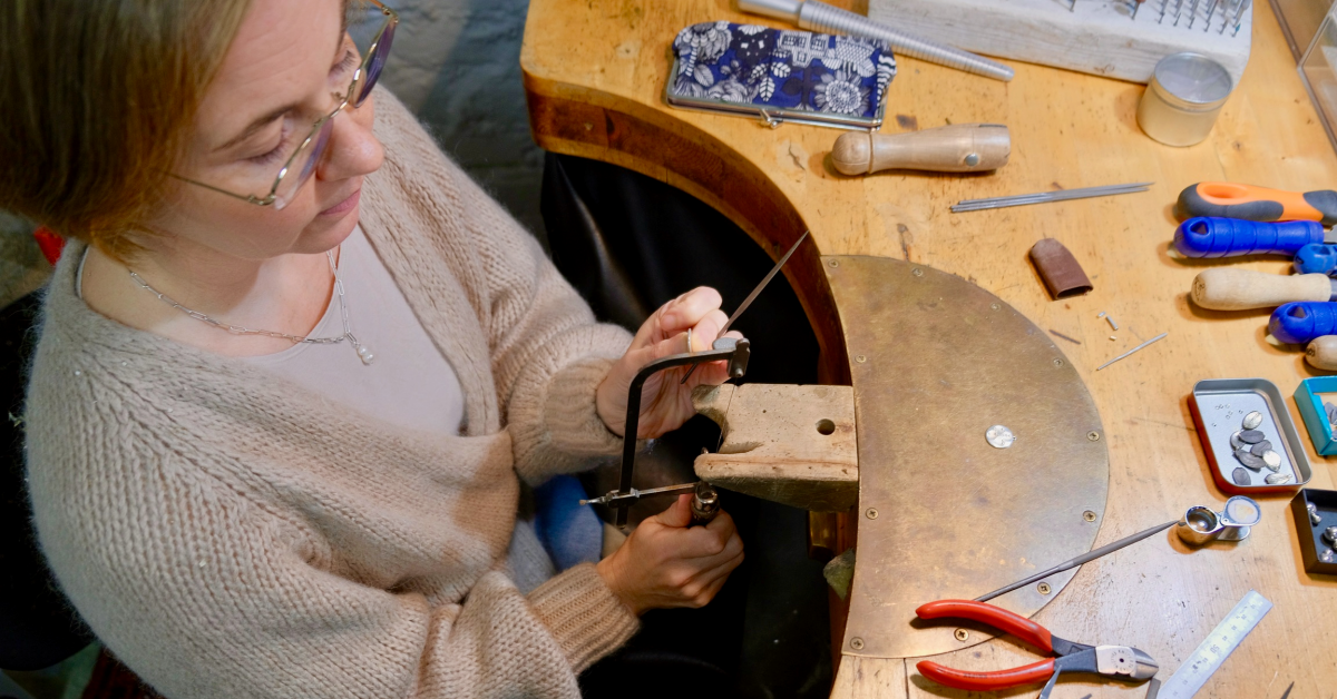 Bildvinkel snett uppifrån. En kvinna med ljust hår och glasögon sitter vid en arbetsbänk och jobbar koncentrerat med en fil och en liten handsåg. På bordsytan framför henne ligger tänger, filar och ett ringverktyg.