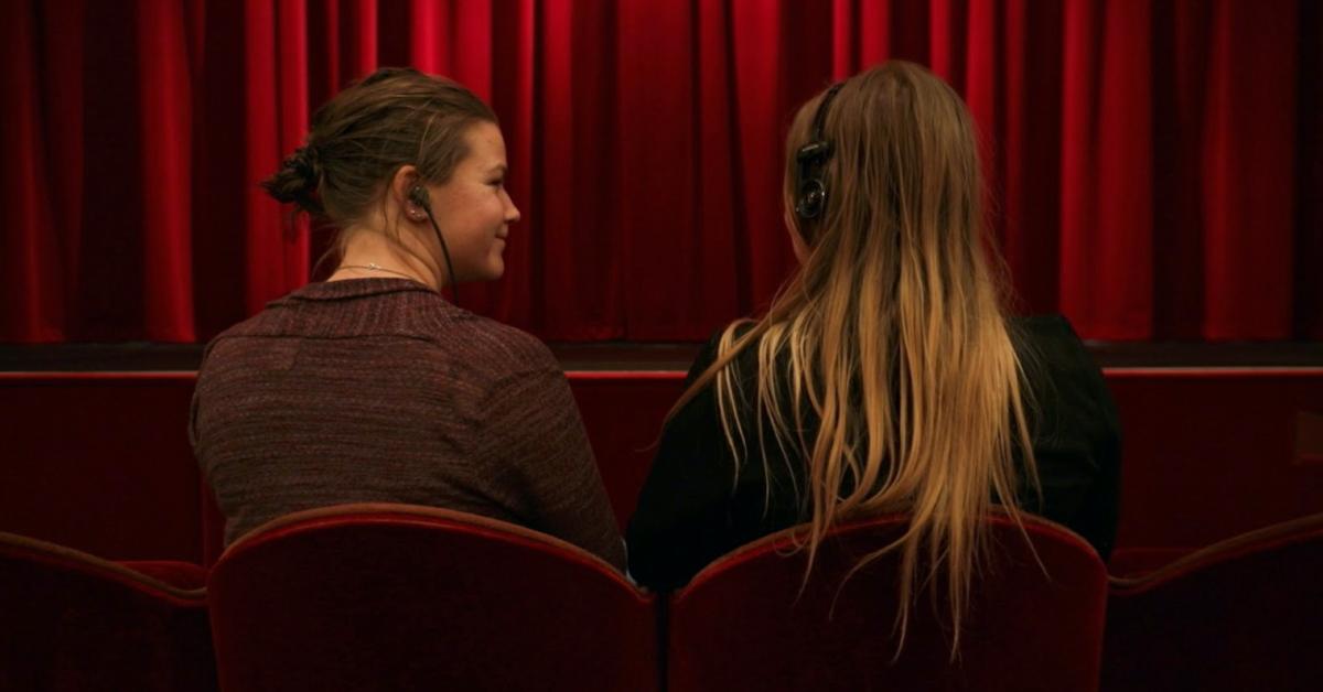 Emma Kullberg och Elin Sundell sitter med syntolkningsutrustning i en teatersalong framför ett rött draperi..