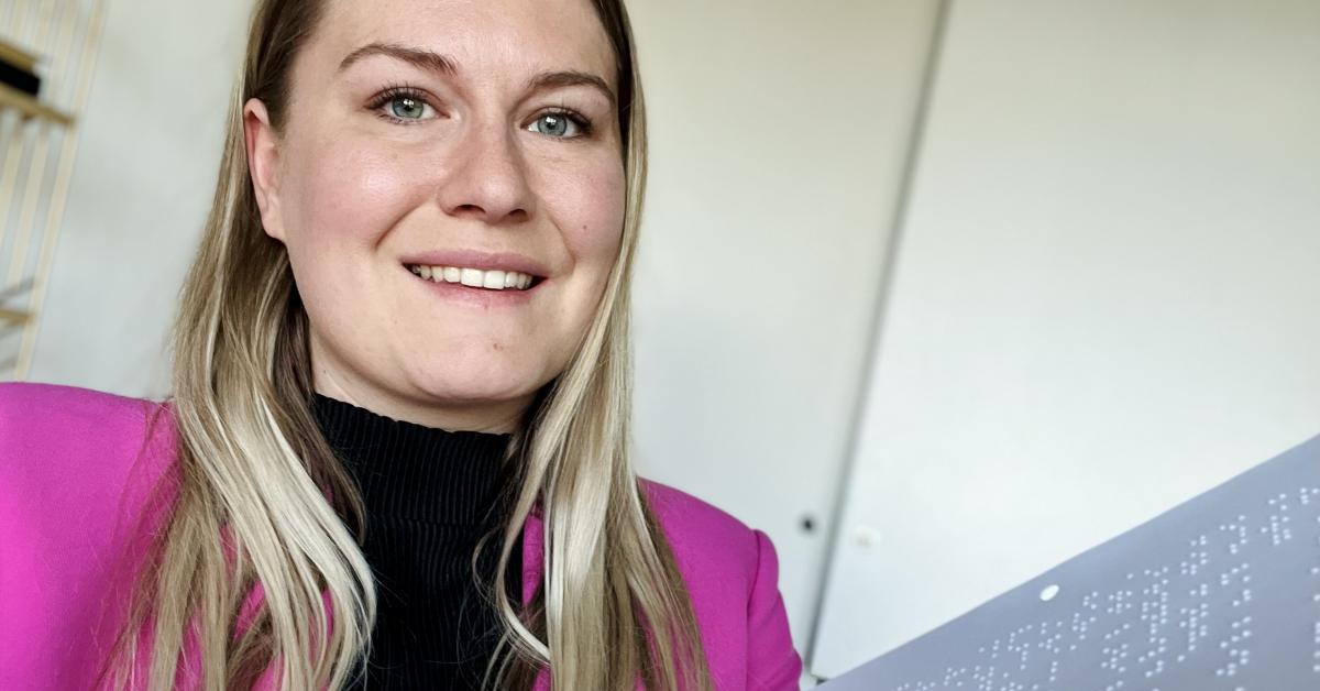 En leende Elin i långt blont hår, svart polotröja och pink kavaj. Elin har en bok i punktskrift i handen.