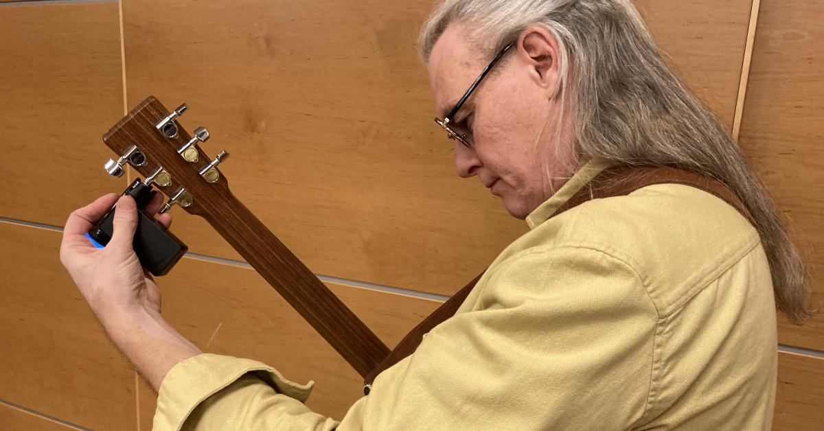 Johan Seige stämmer sin akustiska gitarr inför uppträdandet på FSS jubileumsfest i Tammerfors i november. Han är fotograferad från vänstra sidan i halvbild. Han har en beige kragskjorta och långt grått hår och mörka glasögon. Gitarrhalsen pekar snett uppåt och han stämmer den med vänster pekfinger och tumme.. 