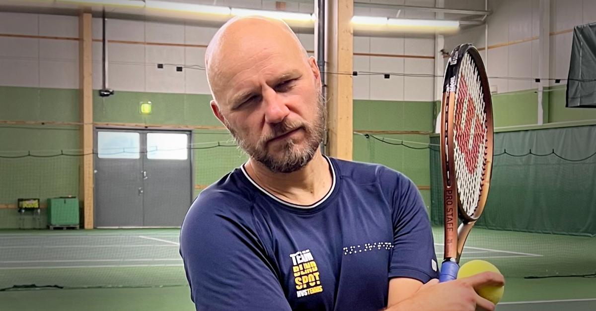 Närbild. En man med kort skägg och rakat huvud står med armarna i kors i en tennishall. Han är klädd i en mörkblå t-skjorta och håller en racket och en tennisboll.