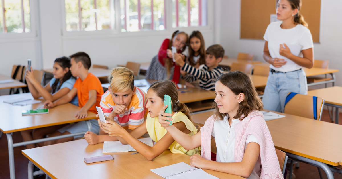 8 skolelever vid pulpeter i ett klassrum fokuserar på sina mobiler. En ung kvinna utan mobil får ingen uppmärksamhet av dem där hon står med papper i händerna.