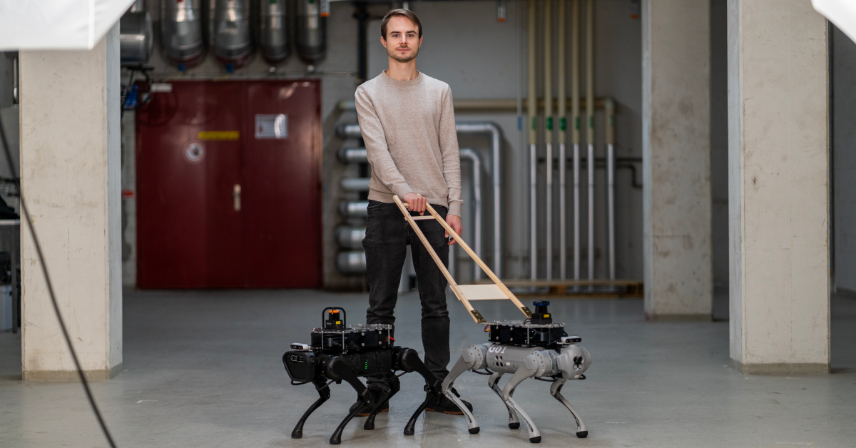 Forskaren Davide Plozza står i sitt laboratorium och visar upp en grå och en svart robotledarhund. Hundarna har böjda ben, kameror och lådliknande elektronik på ryggen. Foto: Marco Giordano