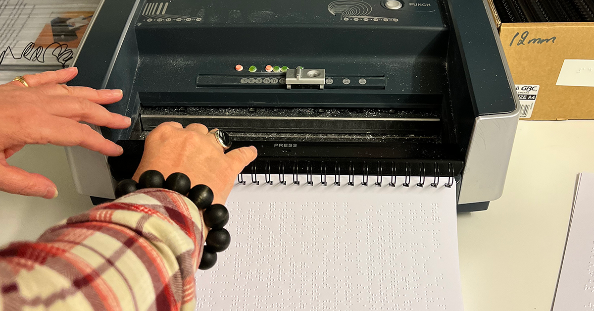 En punktskriftstidning blir till. En närbild av två händer och en punktskriftmaskin med en halvfärdig tidning.