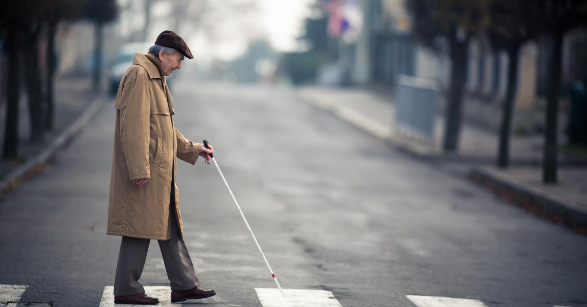 En äldre man går över en skyddsväg med vita käppen. Mannen har en ljus trenchcoat på sig och hatt. I bakgrunden syns hus på båda sidorna om vägen. 