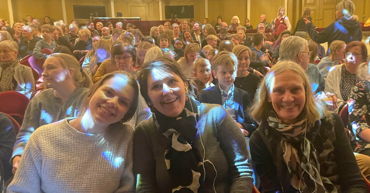 Karen Lindholm, Veera Florica Rajala och Henrika Mercadante sitter i en fullsatt teatersalong och ler mot kameran.