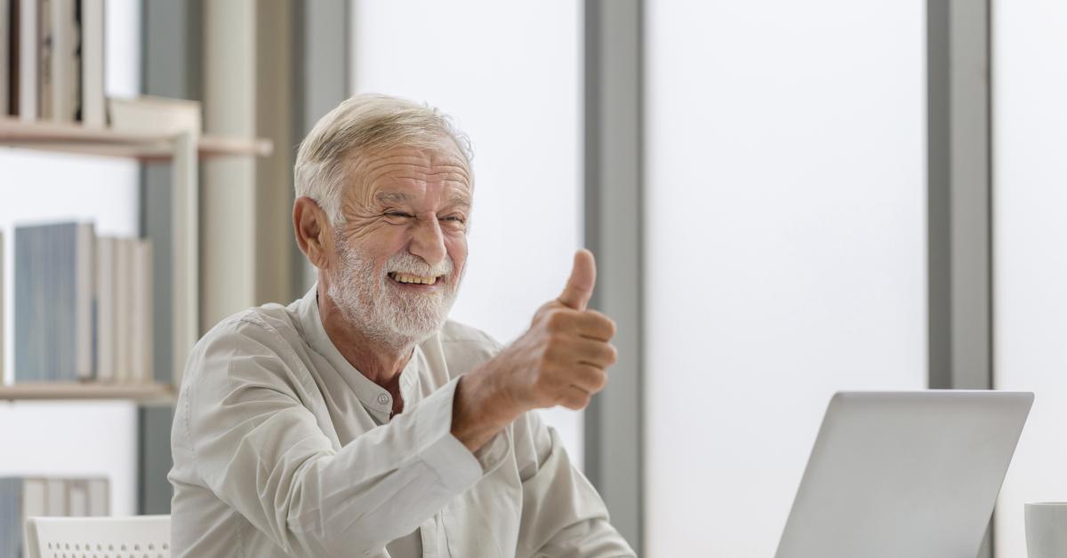 En brett leende äldre vithårig herre med skägg sitter vid en bärbar dator i ett ljust arbetsrum och håller upp tummen. 