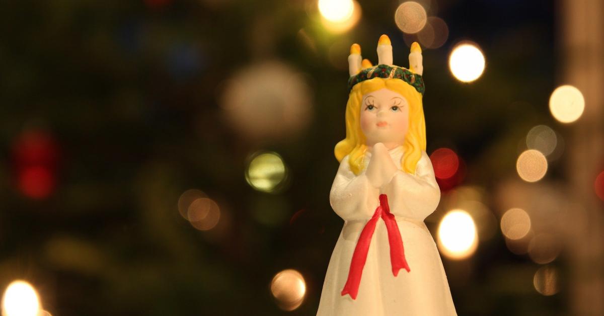 En dekorationsfigur av ljudhårig Lucia med blont hår och ljuskrans och vit klänning med rött skärp. En suddig julgran syns i bakgrunden.