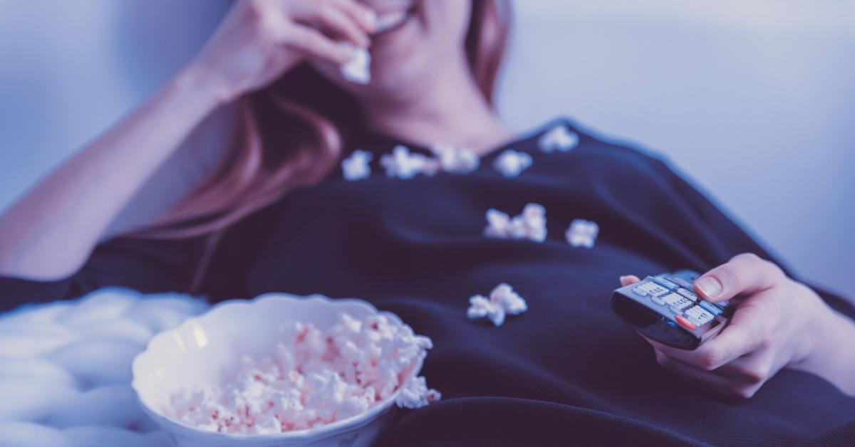 En kvinna, med suddiga konturer, ligger på en soffa och äter popcorn och har en fjärrkontroll i handen.