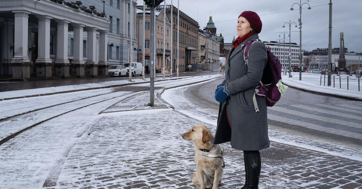 Veera Florica Rajala står med ledarhunden Palo vid en övergång på Salutorget i Helsingfors i vintrigt landskap. Tillsammans med FSS-distrikten och övriga FSS-anställda jobbar hon med påverkan av makthavarna i den nya riksdagen, i städerna och i vårddistrikten.