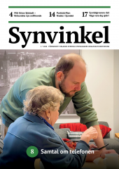 En äldre kvinna med kort, lockigt grått hår och mörkblå kavaj sitter vid ett bord. En ung, skäggig man klädd i grön tröja lutar sig framåt och visar kvinnan något på en mobiltelefon. De befinner sig i ett ljust rum med en grå soffa och röda kuddar i bakgrunden. 