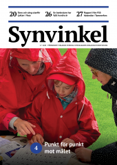 En kvinna och ett barn klädda i röda regnrockar med huva studerar en lapp i punktskrift som ligger intill ett punktskriftsalfabet på ett bord på en sportplan med grönt gräs. En kvinna i svart regnrock med vita prickar ser på.