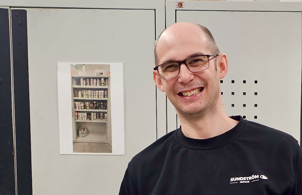 En leende man med glasögon står i arbetskläder framför gråa metallskåp i en lagerlokal. Flera av skåpen har svarta lodräta tejpremsor fästa på kanterna för att markera dem tydligare.