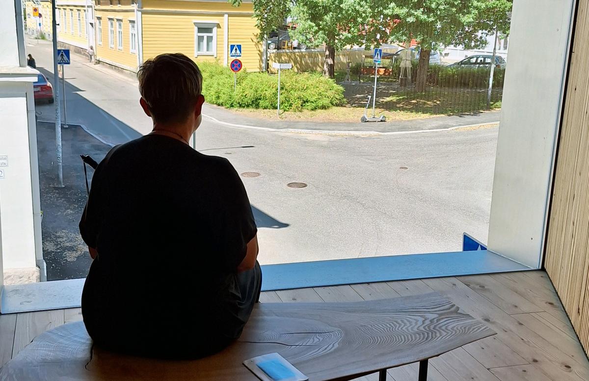 En person med kort hår sitter på en bänk framför ett stort fönster med utsikt över en gata och flera trähus i gul färg. Rummet är inrett i trä med både väggar och golv i samma träton, vilket skapar en varm känsla i interiören.