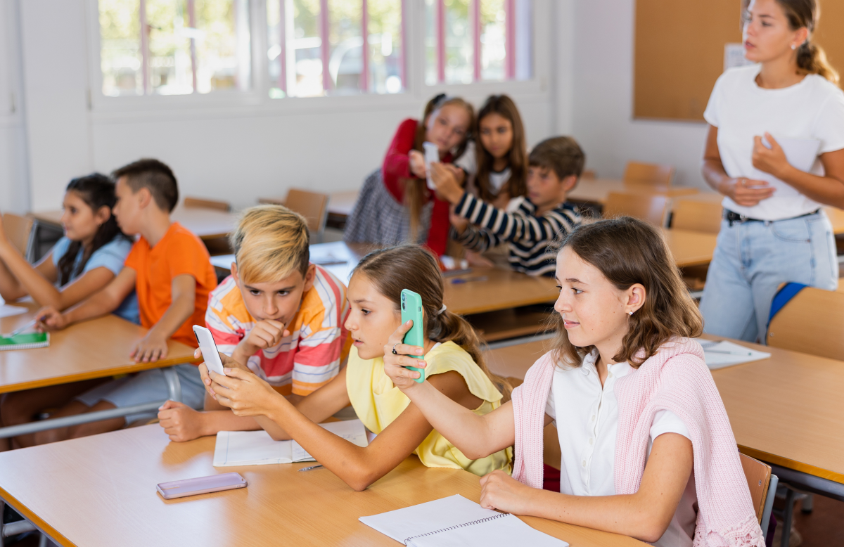 8 skolelever vid pulpeter i ett klassrum fokuserar på sina mobiler. En ung kvinna utan mobil får ingen uppmärksamhet av dem där hon står med papper i händerna.