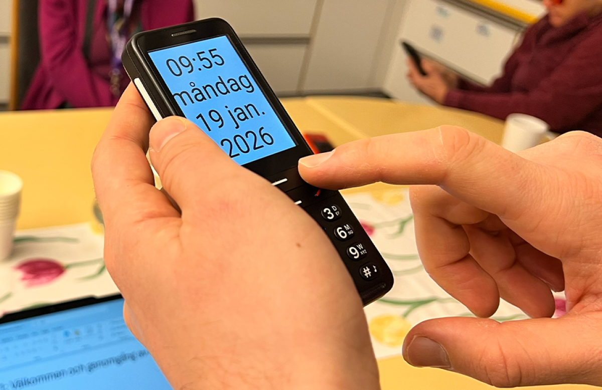 Närbild bakifrån. En person håller upp en svart, avlång mobiltelefon med knappsats och trycker med pekfingret på en knapp. På den stora blåtonade skärmen står klockslaget 09:55 och datumet måndag 19 jan. 2026 med stora tecken. I den oskarpa bakgrunden syns delvis två personer sittande vid ett bord.