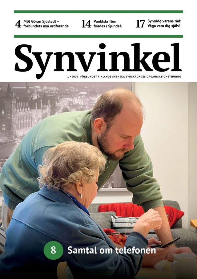 En äldre kvinna med kort, lockigt grått hår och mörkblå kavaj sitter vid ett bord. En ung, skäggig man klädd i grön tröja lutar sig framåt och visar kvinnan något på en mobiltelefon. De befinner sig i ett ljust rum med en grå soffa och röda kuddar i bakgrunden. 